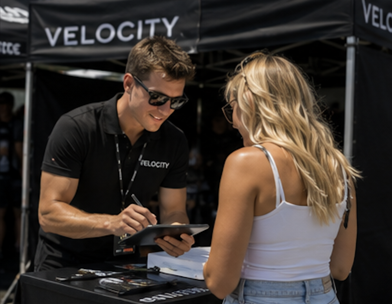 Male event staff member in a black branded polo checking in a guest on a tablet at an outdoor activation booth