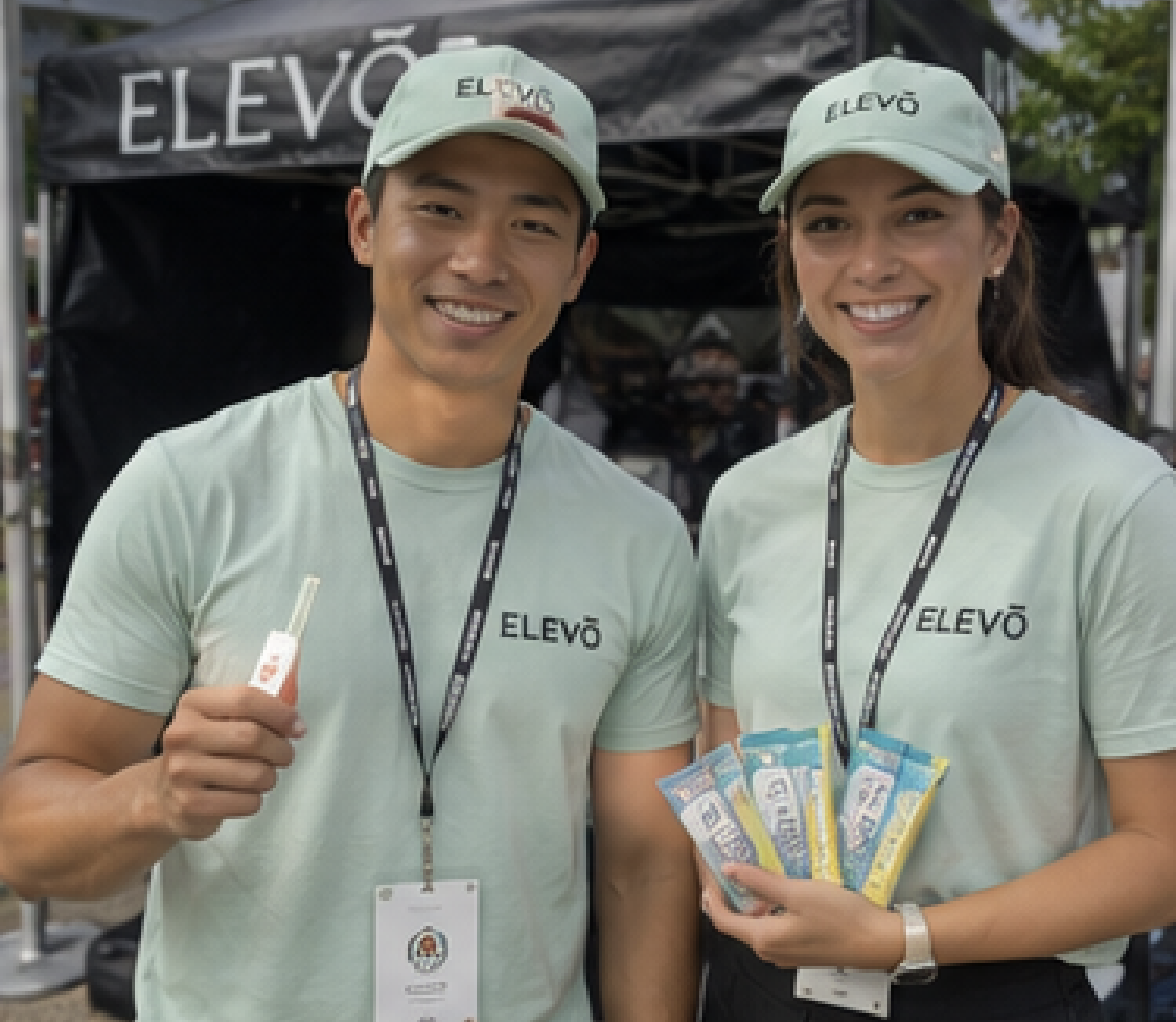 Two promotional staff in mint branded caps and shirts holding product samples in front of a black event tent