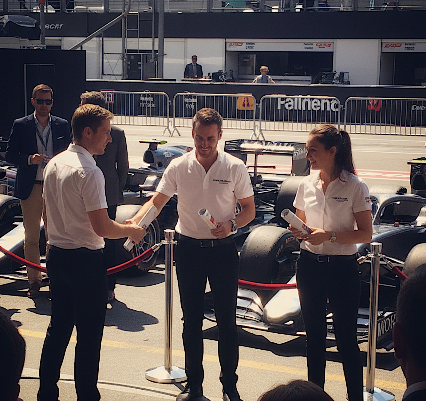 Brand ambassadors in white shirts in the pit lane beside formula racing cars