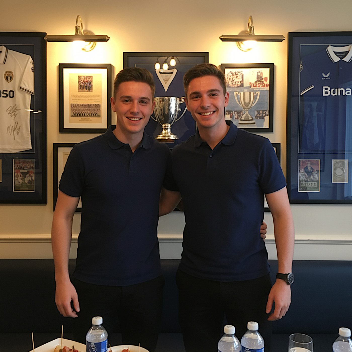 Two hospitality team members in navy polos standing in front of framed memorabilia and a trophy display