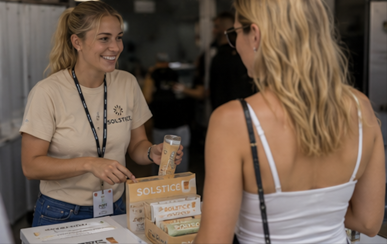 Brand ambassador in a beige branded T-shirt handing product samples to a guest at an expo booth